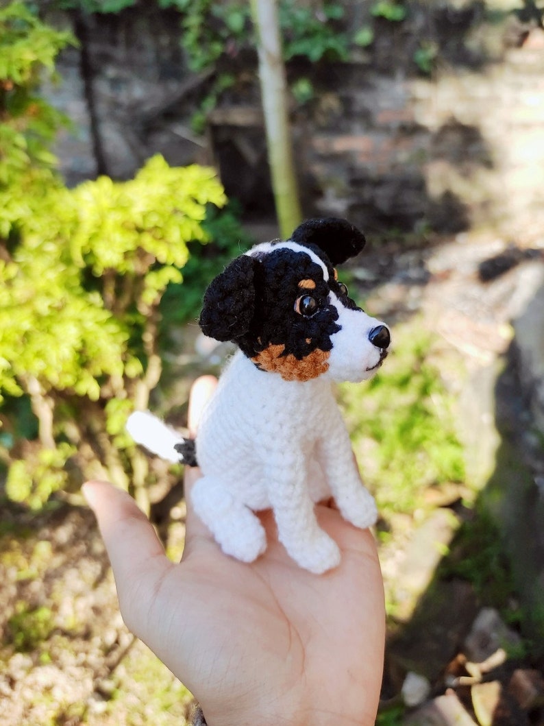 May include: A crocheted dog toy in white, black, and brown. The dog is sitting up with its head tilted to the side. The toy is sitting on a person's hand.
