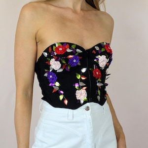Puede incluir: Un corsé negro sin tirantes con bordado floral. Las flores están hechas de tela y lentejuelas en tonos rojos, rosas, morados y verdes.