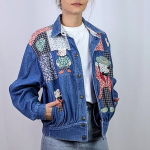 Puede incluir: Una chaqueta vaquera azul con detalles de patchwork que presenta un estampado floral y figuras aplicadas. La chaqueta tiene un cierre de botones en la parte delantera y dos bolsillos.