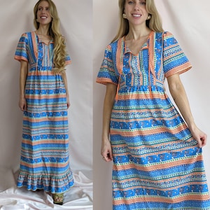 Op de afbeelding: Een vintage maxi-jurk in blauw, oranje en wit. De jurk heeft een V-hals met oranje afwerking en korte mouwen. De stof is bedrukt met strepen en bloemmotieven en de jurk heeft een geplooid zoom.