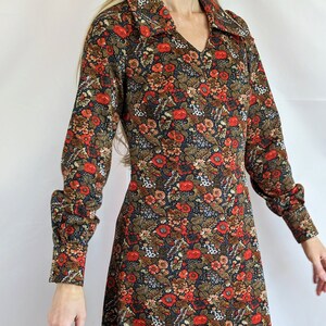 Puede incluir: Un vestido de manga larga con estampado floral en tonos rojos, marrones y blancos sobre un fondo azul oscuro. El vestido tiene cuello y mangas largas con puños abotonados.