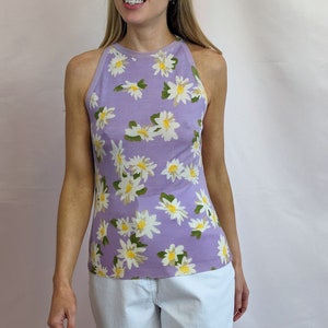 Puede incluir: Un top sin mangas de color lavanda con estampado de margaritas blancas. El top tiene un escote redondo y un corte holgado.