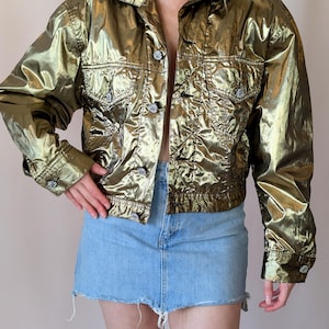 Puede incluir: Chaqueta dorada metálica con botones y dos bolsillos en el pecho. La chaqueta tiene una textura brillante y arrugada y mangas largas. La modelo también lleva una minifalda vaquera azul claro.