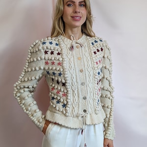 Puede incluir: Un cárdigan de punto color crema con un patrón de punto trenzado y flores bordadas en tonos azules, rosas y rojos. El cárdigan tiene una cintura con cordón y detalles de pompones en las mangas.