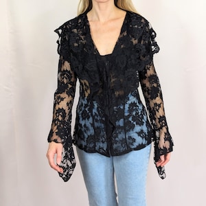 Puede incluir: Una blusa negra de encaje con mangas largas y un cuello con volantes. La blusa es transparente y tiene un estampado floral.