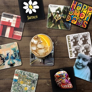 May include: A selection of square drinks coasters displaying album artwork and band names such as "The Stone Roses," "Oasis," and "The Smiths." A glass of iced drink with an orange slice rests on a coaster. The coasters are arranged on a wooden surface.