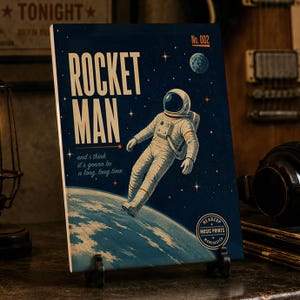 May include: A rectangular art print featuring a dark blue space background with a white astronaut floating above a blue planet. The text reads ROCKET MAN, No. 002, and i think it's gonna be a long, long time. A circular logo reads HEADZUP MUSIC PRINTS MANCHESTER.