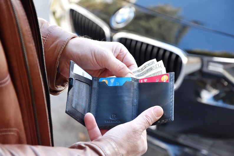 May include: A black leather trifold wallet with multiple card slots and a cash compartment. The wallet is open and shows a few credit cards and some cash.