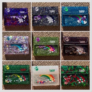 May include: Nine different glitter resin wallets with a rainbow, clouds, and hearts design. Each wallet has a different color and glitter combination. The text "Trapper Keeper" is printed on each wallet.