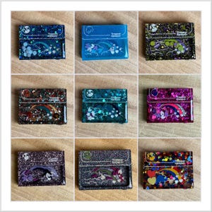 May include: Nine glittery resin cardholders in various colors, each featuring a design with a rainbow, clouds, and the text "Trooper Keeper." The cardholders have a rectangular shape with a flap closure.