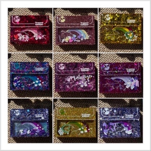 May include: Nine glittery Trapper Keeper-style cases in various colors, each with a clear window showcasing a unique design. The designs include rainbows, clouds, and various decorative elements. The cases are rectangular with a rounded top.