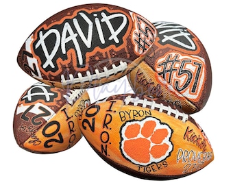 May include: Three brown leather footballs featuring custom hand-painted designs. One displays the name DAVID and number 51. Another shows the number 51. The third features a paw print logo, the text BYRON, IRON, TIGERS, and PROVERBS 27:17.