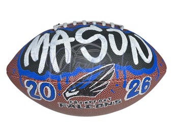 May include: A brown leather football featuring white laces and a custom design. The name MASON is painted in large white letters over a black and blue drip pattern. The numbers 20 and 26 flank a black and blue falcon logo above the text FRANKFORT FALCONS.