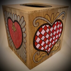 Può includere: Scatola di fazzoletti di legno con disegni di cuori dipinti a mano. La scatola quadrata ha un'apertura superiore. Due lati presentano cuori rossi con bordi decorativi. Un cuore è pieno, l'altro è a scacchiera rossa e bianca.