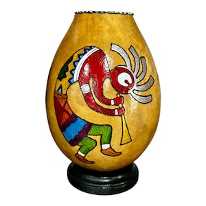 May include: Hand-painted gourd art featuring a Kokopelli figure in red, yellow, blue, and green. The gourd is mounted on a black base. The Kokopelli is depicted playing a flute, with a distinctive headdress. The gourd has a natural, textured surface.