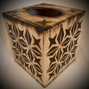 May include: A wooden tissue box with a square shape and a central opening. The box features a light wood finish with dark, geometric starburst patterns carved into the sides. The edges are slightly charred, adding a rustic touch.