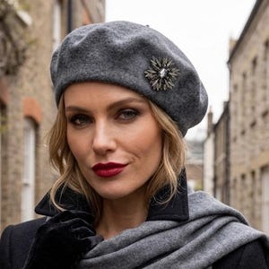 May include: A grey wool beret featuring a silver-toned starburst brooch with a central crystal, paired with a matching grey wool scarf, a black coat with a high collar, and black velvet gloves, creating a coordinated cold-weather ensemble.