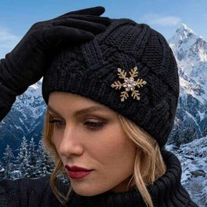May include: A black knit beanie featuring a textured cable pattern and a gold-toned snowflake-shaped brooch with clear crystals. The beanie is paired with a matching black knit glove and a black turtleneck sweater.
