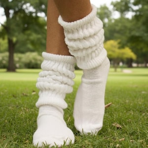Puede incluir: Un par de calentadores y calcetines blancos. Los calentadores son gruesos y acanalados, con un aspecto arrugado. Los calcetines son de color blanco liso. La imagen está tomada al aire libre en un césped.
