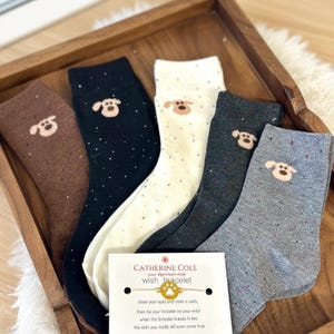 Könnte beinhalten: Fünf Paar Socken in verschiedenen Farben, darunter Braun, Schwarz, Weiß, Dunkelgrau und Hellgrau. Jede Socke hat ein kleines Hunde-Gesicht-Design. Eine "Catherine Cole" Wunscharmband-Karte ist ebenfalls vorhanden.