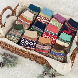 May include: A wicker basket filled with a variety of colorful patterned socks. The socks feature geometric and traditional designs in shades of blue, green, red, and brown. The basket sits on a white, fluffy surface, with pinecones and greenery adding a seasonal touch.