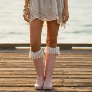 May include: Light pink knee-high rubber boots featuring white lace trim at the top cuffs. The boots are worn with a light-colored, semi-sheer tunic dress that has a subtle embroidered pattern along the hemline and short sleeves.