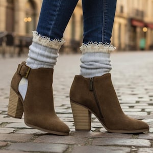 May include: Brown suede ankle boots with a block heel and side zip. The boots feature a buckle detail and a cutout design. The person is wearing blue jeans and grey socks with a lace trim.