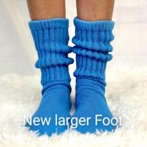 May include: A pair of bright blue slouch socks with the text "New larger Foot" at the bottom. The socks are made of a soft, ribbed knit material and are gathered around the ankles. The socks are displayed on a white, fluffy surface.