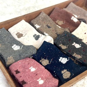 Könnte beinhalten: Eine Holzkiste enthält eine Sammlung von Socken mit Muster. Die Socken sind in verschiedenen Farben, darunter Grau, Marineblau und Rosa, mit einer gesprenkelten Textur. Jede Socke hat ein sich wiederholendes Katzensilhouetten-Design in Weiß oder Braun.