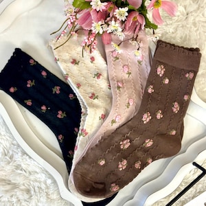 May include: Four pairs of socks in varying colors: navy, cream, pink, and brown. Each sock features a floral pattern with pink roses. The socks are arranged on a white tray with a floral arrangement.