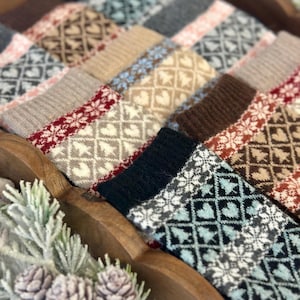 May include: A collection of patterned socks in various colors, including gray, brown, and black. The socks feature geometric and snowflake designs. The socks are arranged in a wooden tray, suggesting a cozy, winter theme.