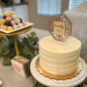 Peut inclure: Un gâteau blanc avec un topper de gâteau doré qui dit "Emily" et "Emily" en coréen. Le gâteau est sur un support à gâteau blanc avec une bordure dorée. Il y a des feuilles vertes et des fleurs blanches autour du support à gâteau. Il y a aussi d'autres friandises sur la table, y compris une assiette de gâteaux de riz colorés.