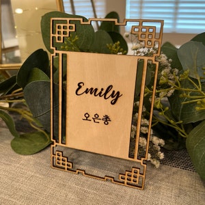 May include: A wooden table number card with a decorative frame. The card is engraved with the name "Emily" and the Korean text "오은총".