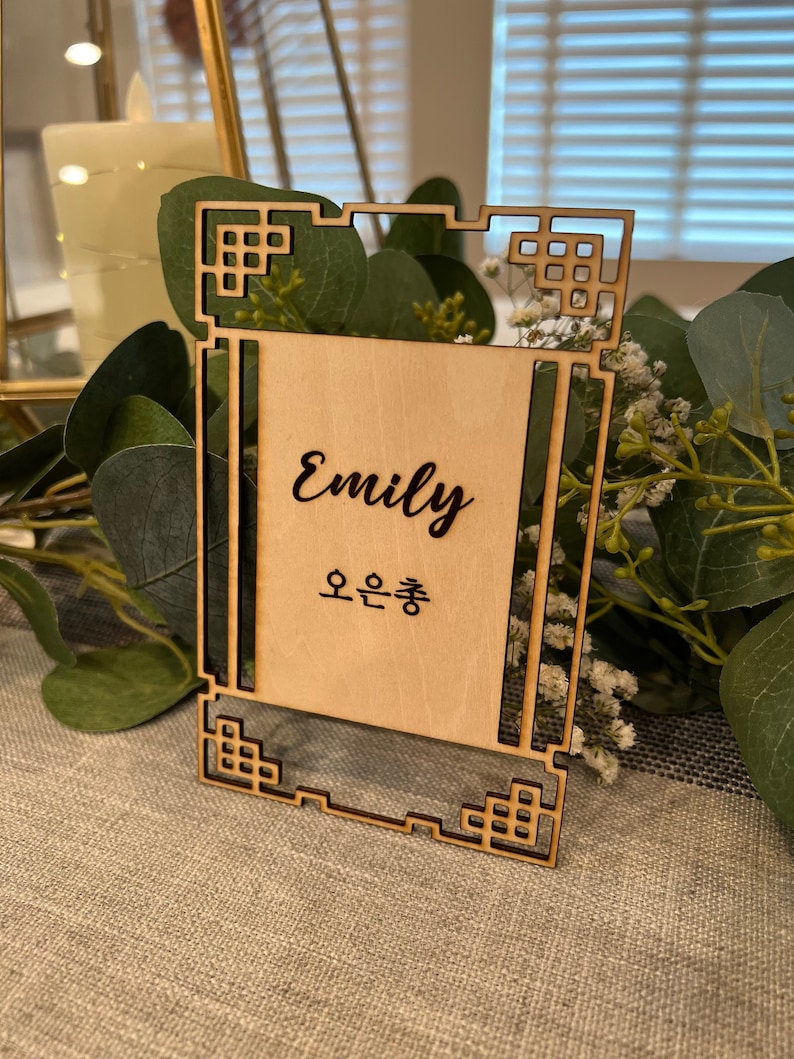 May include: A wooden table number card with a decorative frame. The card is engraved with the name "Emily" and the Korean text "오은총".