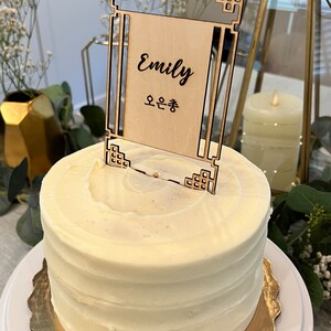 May include: A white cake with white frosting and a wooden cake topper with the name "Emily" and the Korean text "오은총" written on it.