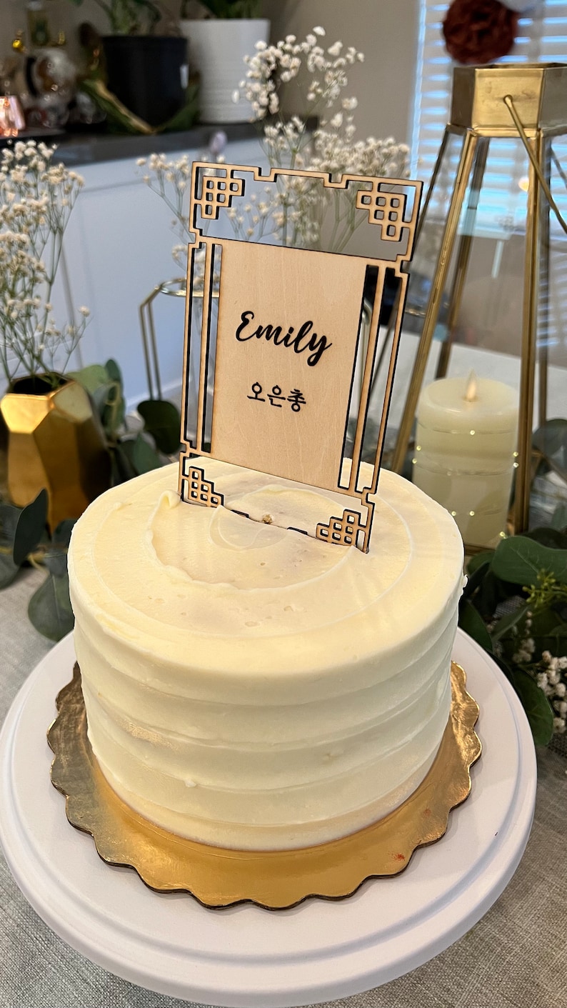 May include: A white cake with white frosting and a wooden cake topper with the name "Emily" and the Korean text "오은총" written on it.
