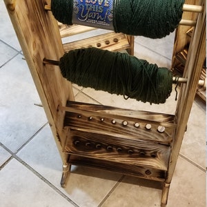May include: A wooden yarn holder with two spools of green yarn. The holder has a shelf with holes for storing knitting needles. The yarn is labelled "I Love This Yarn!"