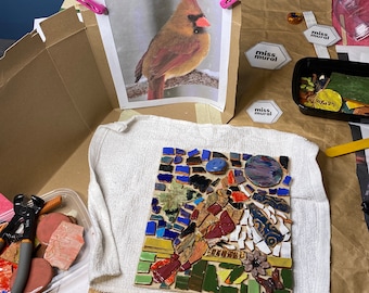 May include: A close-up of a mosaic art project in progress. The mosaic is made of small, colorful tiles and is set on a white towel. The mosaic depicts a cardinal bird, a popular motif in art and nature.
