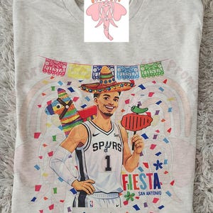 May include: A light gray t-shirt featuring a colorful graphic of a basketball player wearing a sombrero, holding a grilled pepper, and a piñata. The shirt has the text "Fiesta San Antonio" and "Spurs 1".