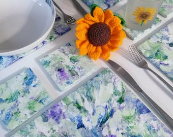 May include: A table setting featuring a white bowl with blue stripes, floral placemats, coasters, and a crocheted sunflower. The placemats and coasters have a blue, green, and purple floral design. A glass with a sunflower design is also present.