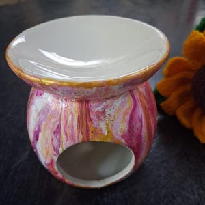 May include: A ceramic wax warmer featuring a marbled paint finish in shades of pink, white, and gold. The piece has a wide, shallow bowl at the top for wax and a circular opening at the base for a tea light candle, with gold paint accents.