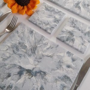 May include: A collection of grey and white marble-effect placemats and coasters. The placemats are rectangular, and the coasters are square. A crocheted sunflower and cutlery are also visible.
