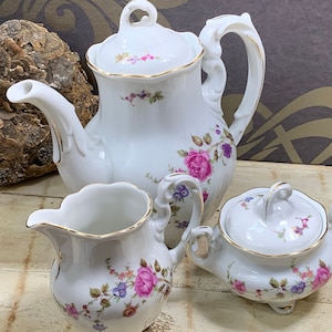 Może przedstawiać: Biały porcelanowy zestaw do herbaty z kwiatowymi wzorami. Zestaw zawiera czajnik, dzbanek do śmietanki i cukiernicę.