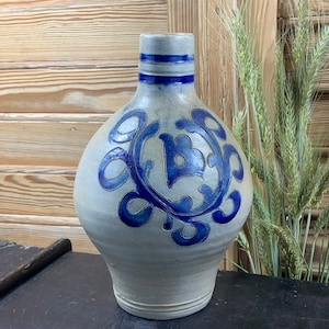 Könnte beinhalten: Eine blaue und weiße Keramikkanne mit einem dekorativen Design. Die Kanne hat einen runden Körper und einen schmalen Hals. Das Design zeigt einen blauen Kreis mit einem stilisierten Buchstaben B in der Mitte.