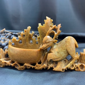 Puede incluir: Una escultura de ciervo tallada a mano en piedra con detalles intrincados. El ciervo sostiene un pequeño cuenco en su boca. La escultura está hecha de piedra marrón claro y tiene un aspecto rústico y antiguo.