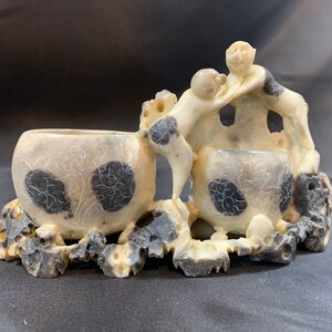 May include: A white and black carved stone sculpture of two monkeys playing with a ball. The monkeys are perched on a rock base with two carved bowls.