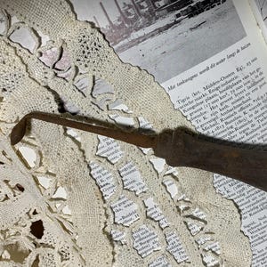 May include: An antique, rusty metal tool with a wooden handle, possibly for leatherwork or a similar craft. The tool is set against a backdrop of an open book and lace.