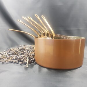 May include: A set of five copper-colored nesting pots with gold-colored handles. The pots are arranged in a stack, with the largest pot at the bottom and the smallest pot at the top.