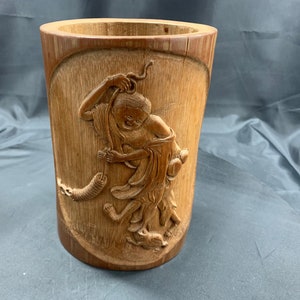 Op de afbeelding: Een cilindrische bamboe vaas met een gesneden ontwerp van een persoon die een lang voorwerp vasthoudt. De vaas is gemaakt van natuurlijke bruine bamboe en heeft een gladde, gepolijste afwerking.