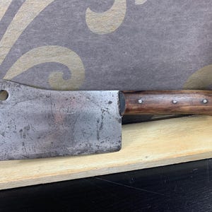 Könnte beinhalten: Ein Vintage-Hackmesser mit einer rechteckigen, abgenutzten Stahlklinge und einem dunklen Holzgriff. Der Griff ist mit drei Nieten befestigt. Das Hackmesser hat ein Loch in der Nähe der Oberseite der Klinge. Das Hackmesser liegt auf einer Holzoberfläche.
